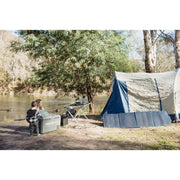 A portable four-panel solar blanket laid out on the ground in a sunny campsite next to a tent. The blue solar panels are angled toward the sun to collect energy for outdoor camping equipment.