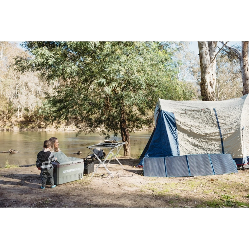 A portable four-panel solar blanket laid out on the ground in a sunny campsite next to a tent. The blue solar panels are angled toward the sun to collect energy for outdoor camping equipment.