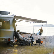 Two people sitting at a table outside an RV with bicycles attached to the back, near a lake.

