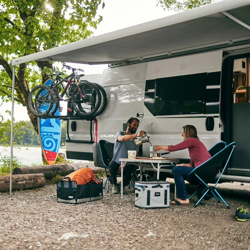 Two people sitting at a table outside an RV with bicycles attached to the back, near a lake.