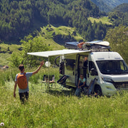 Two people by a white van in a grassy field with mountains in the background