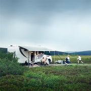 A white motorhome with a large awning extended, parked in a grassy field. A family is sitting outside under the awning, and children are playing nearby.
