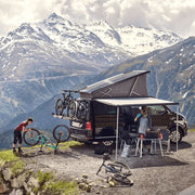 A dark camper van with a Thule 4200 Awning extended, parked in the mountains. Two people are setting up camp with bikes and gear.