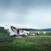 A white motorhome with a large awning extended, parked in a grassy field. A family is sitting outside under the awning, and children are playing nearby.