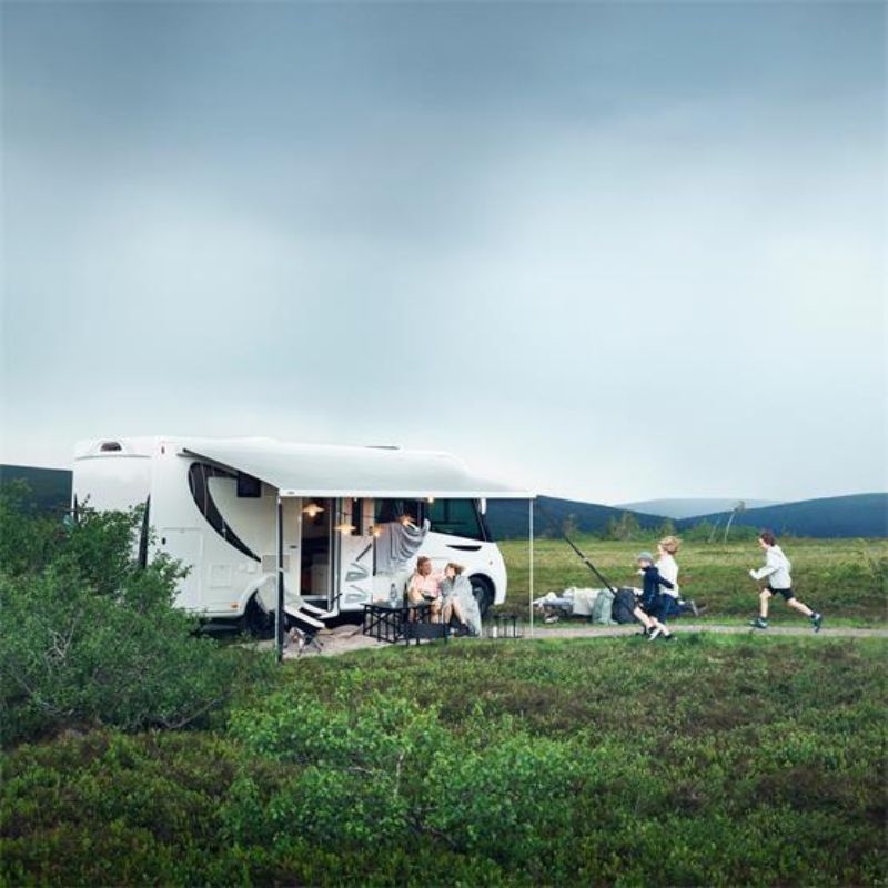 A white motorhome with a large awning extended, parked in a grassy field. A family is sitting outside under the awning, and children are playing nearby.