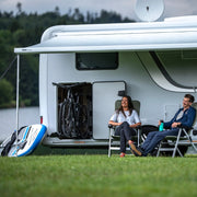 Two people by a white van in a grassy field with mountains in the background
