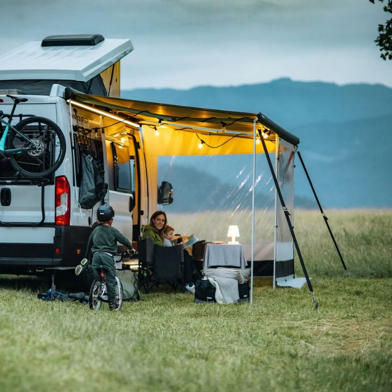Three people by a white van in a grassy field with mountains in the background