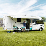 Camper enjoying an outdoor meal set up under an extended awning of a sleek white motorhome on a sunny day.

