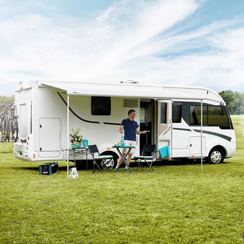 Camper enjoying an outdoor meal set up under an extended awning of a sleek white motorhome on a sunny day.

