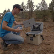 A man in a blue shirt and black cap is shown in a rocky outdoor setting using the icebox, highlighting its internal tray and storage capabilities.