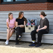 A rugged, light-colored icebox/cooler is open on a bench, displaying its interior storage and a selection of beverages and food, with three young adults seated around it.