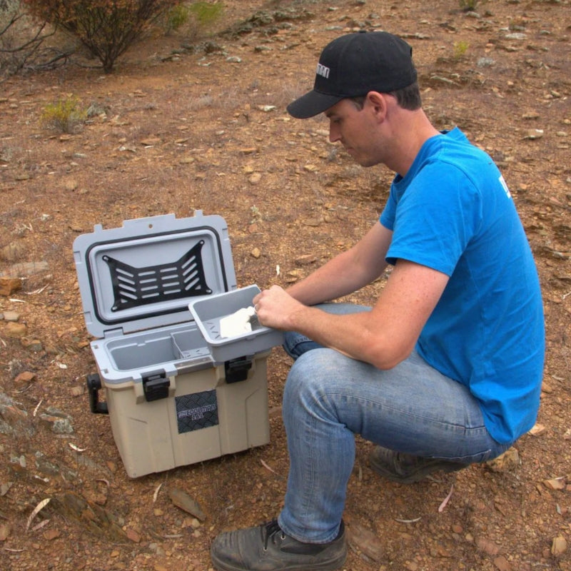 A man in a blue shirt crouches next to an open, rugged tan icebox on rocky ground. The icebox features a grey interior with a removable top tray and an integrated black storage net on the underside of the lid.