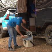 A rugged, tan portable icebox with a grey interior and heavy-duty black latches. The open lid features a black mesh storage net on the underside and a removable grey internal tray for organized food storage.