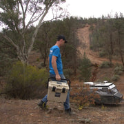 A rugged, tan-colored icebox with a grey interior, featuring a lid-mounted storage net and a removable grey internal tray. The open cooler shows organized compartments for snacks and beverages.