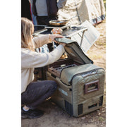 A myCOOLMAN 55 Dual Zone fridge with a khaki Recreational Series cover sits on a riverbank connected to a power station.