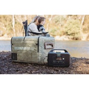 A myCOOLMAN 55 Dual Zone portable fridge fitted with a khaki Recreational Series insulated cover, sitting on a rocky riverbank next to a portable power station.