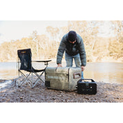 A man adjusts a myCOOLMAN 55 Dual Zone fridge protected by a khaki insulated cover. The unit is powered by a portable battery station on a rocky riverbank at a campsite.