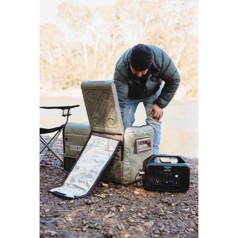 A man adjusts a myCOOLMAN 55 Dual Zone fridge protected by a khaki insulated cover. The unit is powered by a portable battery station on a rocky riverbank at a campsite.