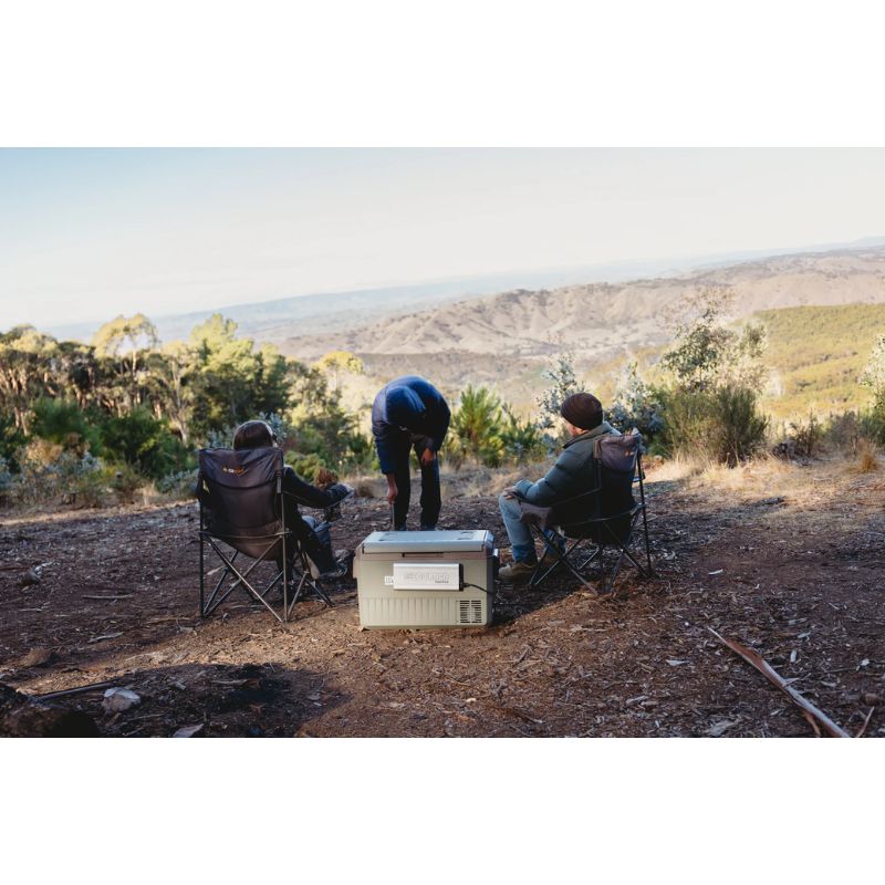 A myCOOLMAN 53L Single Zone portable fridge sits on a rocky riverbank connected to a portable power station. The unit is shown in use at a scenic campsite, providing reliable cooling for a family and a man by the water.