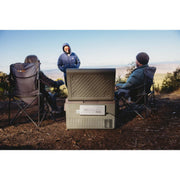 A myCOOLMAN Single Zone portable fridge sits on a rocky riverbank powered by a portable power station. The unit is shown in use by a family and a man at a scenic campsite by the water.