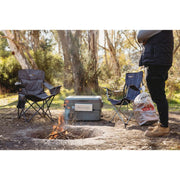 A myCOOLMAN 53L Single Zone portable fridge sits on a rocky riverbank powered by an external battery pack. The outdoor scene shows the unit being used by a family and a man at a scenic campsite by a river.