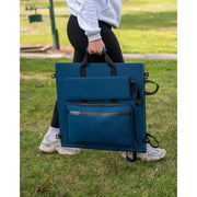 Close-up of a person carrying a folded blue solar blanket by its integrated handle. The folded unit shows its durable fabric exterior, black carrying straps, and a zippered storage pocket for cables or accessories